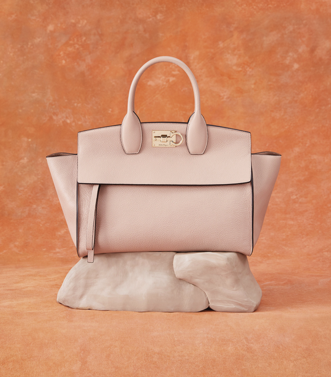 A pale pink Ferragamo handbag with a structured shape and a gold clasp closure. The bag is placed on a smooth, stone-like surface against a warm, textured orange backdrop, emphasizing its elegant and refined design.